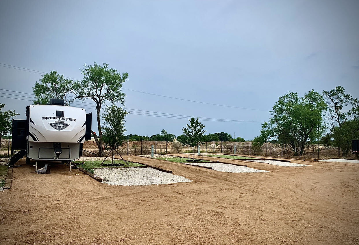 A fifth wheel on its gravel pad with three empty landscaped RV lots stretching toward open Hill Country beyond the fence line