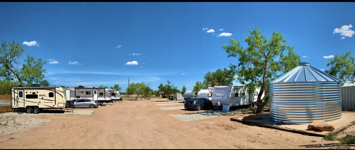 Wide view of the Z-Ranch RV grounds: travel trailers parked along a caliche drive beside mesquite trees and the galvanized grain-bin gazebo